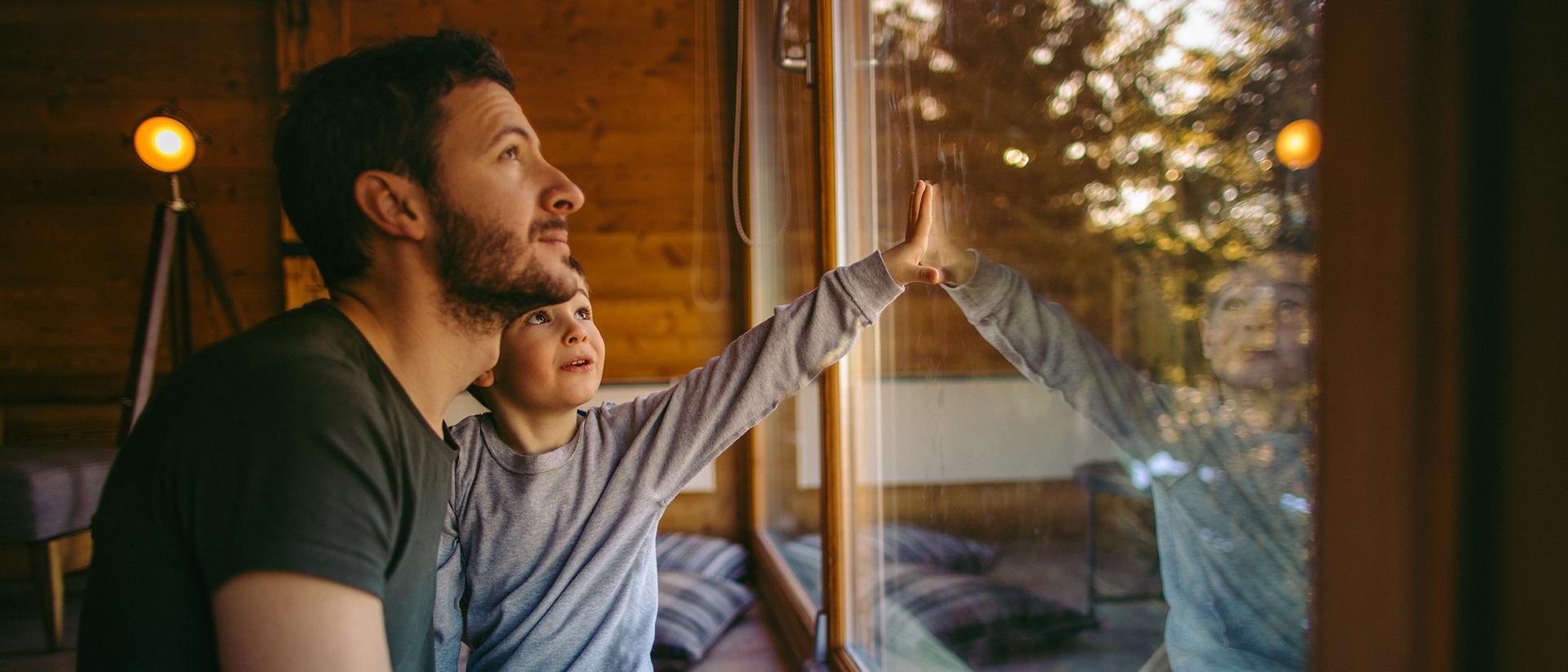 Person and child gaze out a window in a wooden cabin, with warm lighting.